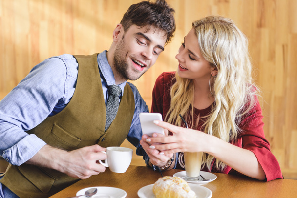 beautiful couple holding coffee cups and using smartphone at cafe  - Foto, afbeelding