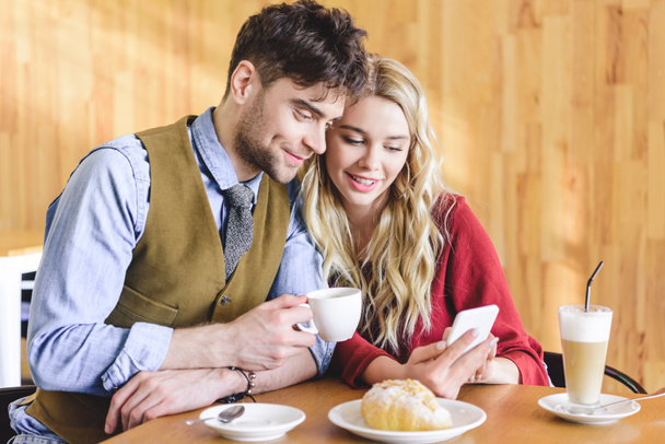 przystojny mężczyzna trzymał filiżankę kawy i kobieta przy użyciu smartfona w kawiarni  - Zdjęcie, obraz