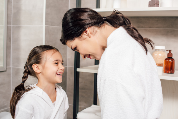 mère regardant à fille souriante en peignoir blanc près des étagères dans la salle de bain
 - Photo, image