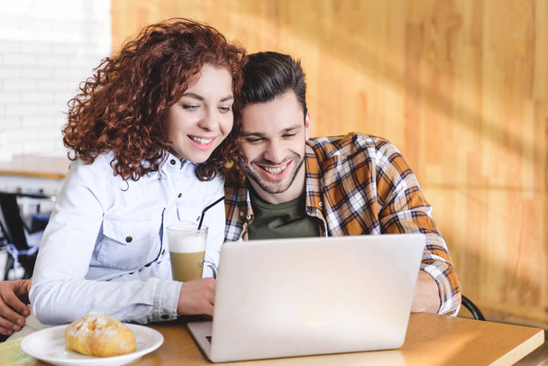 beautiful and smiling couple hugging and using laptop at cafe  - Foto, Imagen