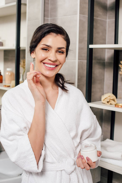 woman in bathrobe holding container and applying cosmetic cream in bathroom - Foto, Imagen