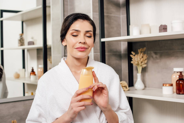 woman in white bathrobe holding bottle with shower gel in bathroom - Photo, Image