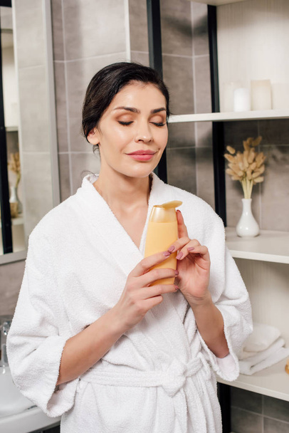 woman in bathrobe holding bottle with shower gel in bathroom - Foto, Bild