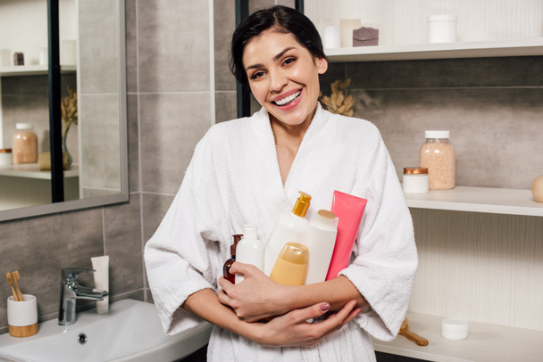 woman in white bathrobe holding bottles and smiling in bathroom - Фото, изображение