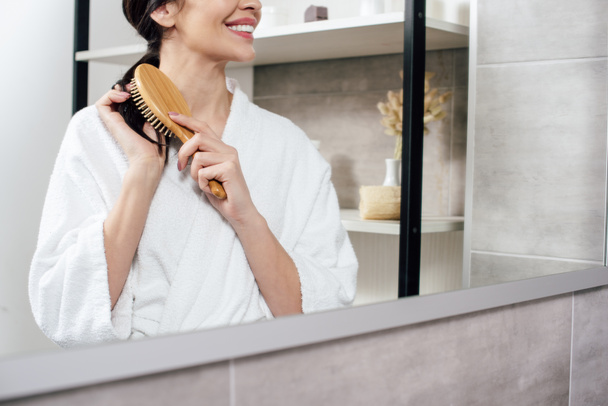 oříznutý pohled ženy v bílém županu česání vlasů a při pohledu do zrcadla v koupelně - Fotografie, Obrázek