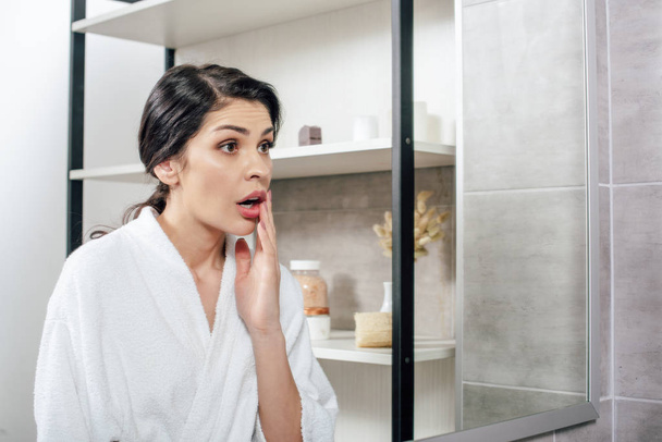 agitated woman in white bathrobe looking to mirror in bathroom - Foto, Bild