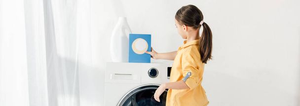 tiro panorâmico da criança de pé perto da máquina de lavar e colocar o pó de lavagem na lavanderia
 - Foto, Imagem
