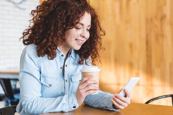 beautiful and smiling woman holding paper cup and using smartphone  - Foto, Bild