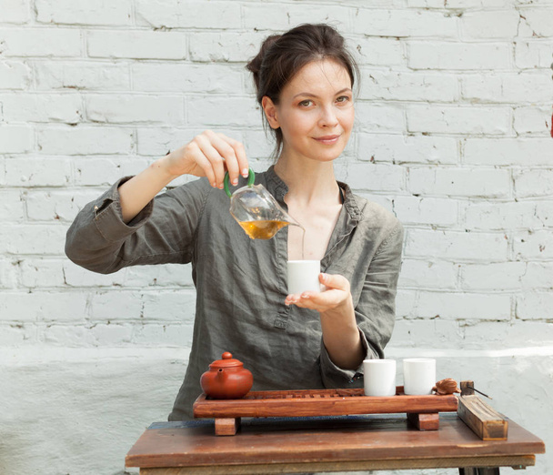Східний майстер чайної церемонії з білою цегляною стіною на фоні. Традиційна чайна вечірка на природі з жінкою, одягнена у сіру сорочку
 - Фото, зображення