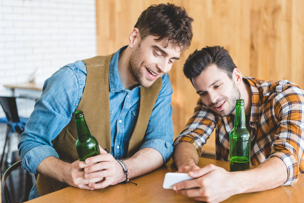 handsome and smiling men holding glass bottles of beer and using smartphone  - Foto, Bild
