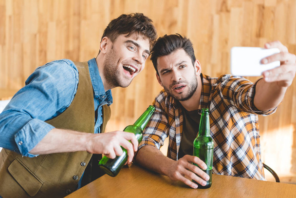 hombres guapos sosteniendo botellas de vidrio de cerveza y tomando selfie en la cafetería
  - Foto, Imagen