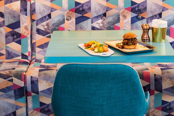 Mesa de restaurante con hamburguesa fresca y ensalada servida en bandejas blancas
 - Foto, imagen