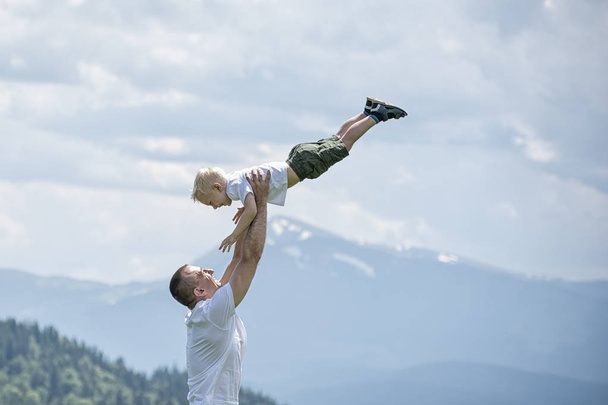 Feliz padre lanza a un pequeño hijo sobre un fondo de bosque verde, montañas y cielo con nubes. Paternidad amistad
 - Foto, imagen