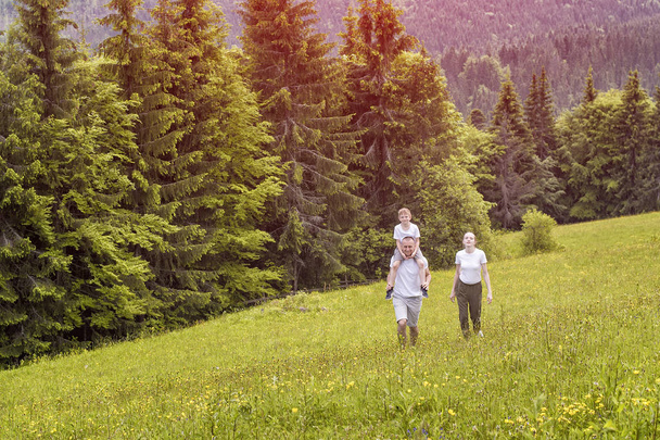 Famille heureuse : père avec fils sur les épaules et mère vont sur un champ vert sur un fond de pinède
 - Photo, image
