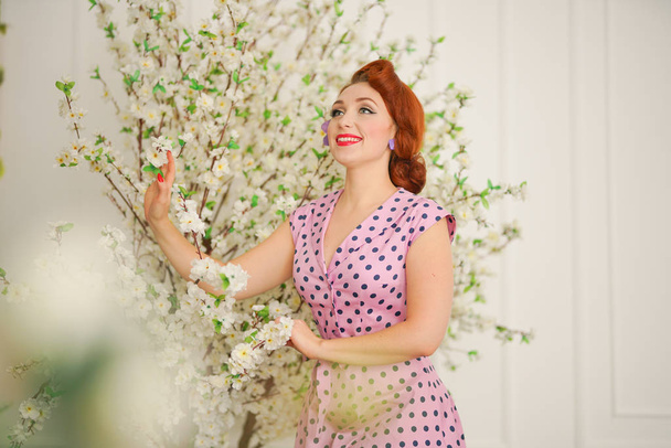 pretty pin up girl in retro polka dot dress near tree with white flowers - Photo, Image