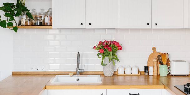Tulips bouquet in vase standing on wooden countertop in the kitchen. Modern white u-shaped kitchen in scandinavian style. - Photo, image