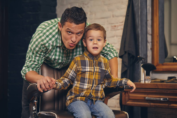 スタイリッシュな小さな子供の背景に若い父親と理髪店の椅子に座って - 写真・画像