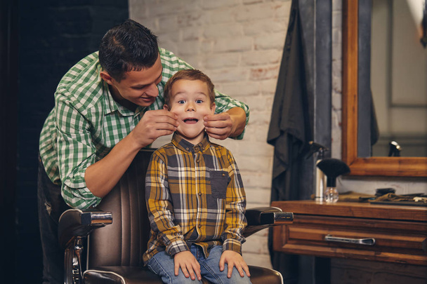 stijlvolle klein kind zit op stoel bij kapsalon met jonge vader op achtergrond - Foto, afbeelding