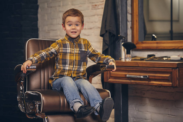Portret van een stijlvolle kleine jongen gekleed in het shirt en spijkerbroek in de barbershop, zittend in een stoel tegen de kappers-werkplek - Foto, afbeelding