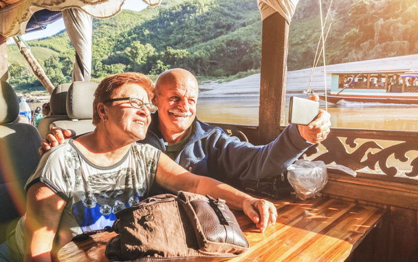 Casal aposentado sênior de turistas felizes tirando selfie na excursão de exploração Mekong com barco lento no Laos PDR - Conceito de viagem idoso ativo em viagem ao redor do mundo - Filtro de pôr do sol saturado quente
 - Foto, Imagem