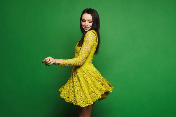 Sexy and fashionable brunette model girl with blue eyes and bright makeup in short stylish yellow dress spin around at green background - Zdjęcie, obraz