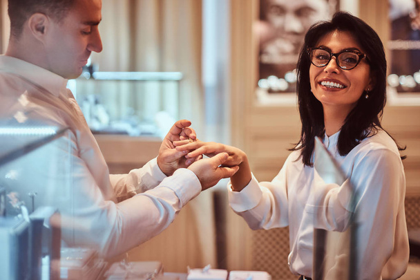 Elegantly dressed man trying on the wedding ring on the hand of his girlfriend in a jewelry store - Foto, Imagem