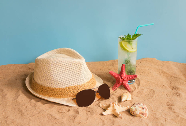 Vaso de cóctel mojito, sombrero, gafas de sol, estrellas de mar y conchas marinas en la playa de arena sobre fondo azul
. - Foto, imagen