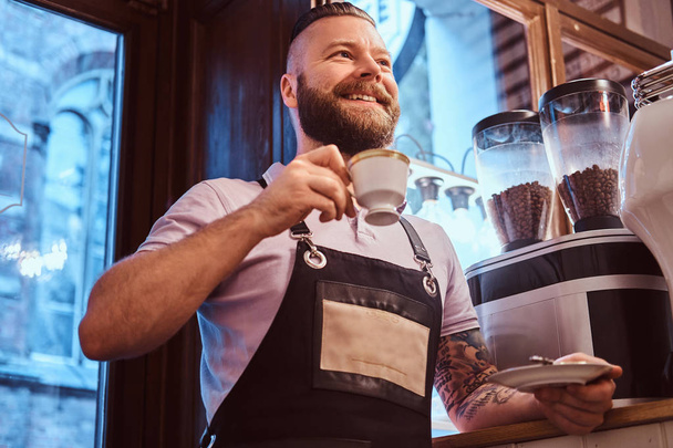 Happy barista em avental segurando uma xícara e um pires, bebendo café durante a pausa para o almoço apoiando-se em um balcão na cafeteria
. - Foto, Imagem