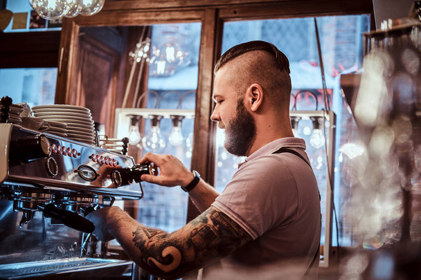 Barista tatuato con barba elegante e acconciatura che lavora su una macchina da caffè in una caffetteria o ristorante
 - Foto, immagini
