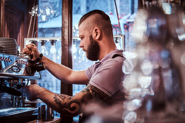 Tattooed barista with stylish beard and hairstyle working on a coffee machine in a coffee shop or restaurant - Foto, Imagem