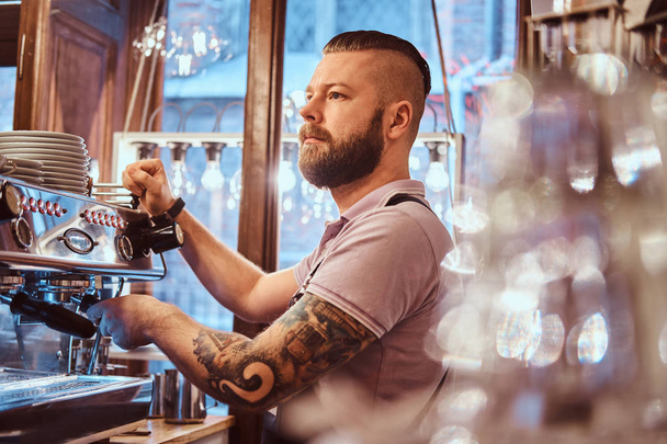 Tattooed barista with stylish beard and hairstyle working on a coffee machine in a coffee shop or restaurant - Photo, Image