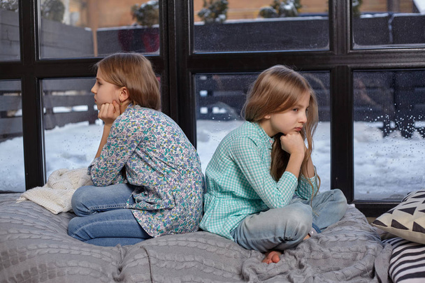 Close up photo of two cute offended little sisters who sit with their backs to each other on windowsill - Photo, Image