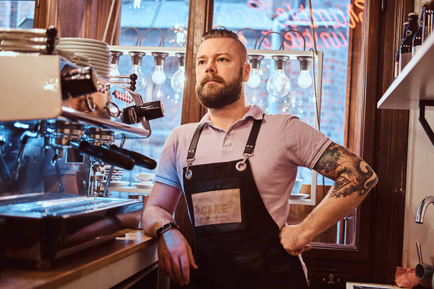 Şık sakal ve saç modeli kafe ya da Restoran bir sayaçta yaslanmış bir kamera için poz önlük giymiş yakışıklı barista - Fotoğraf, Görsel