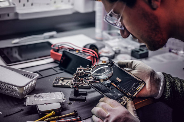 Technicien utilise une loupe pour inspecter soigneusement les parties internes du smartphone dans un atelier de réparation moderne
 - Photo, image