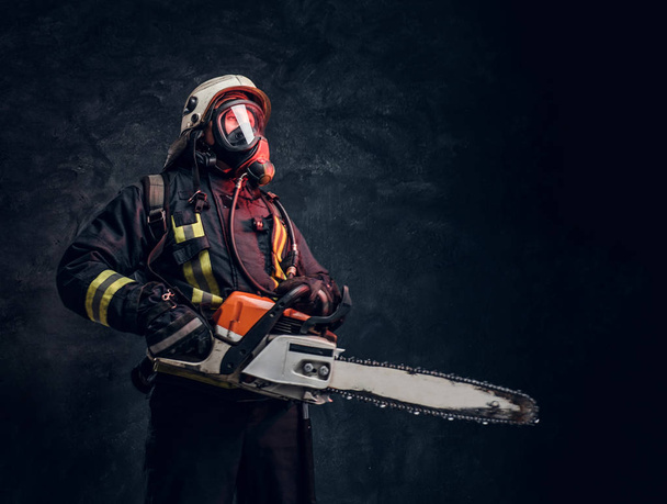 Portrait d'un pompier portant un casque de sécurité et un masque à oxygène tenant une tronçonneuse. Studio photo contre un mur sombre texturé
 - Photo, image