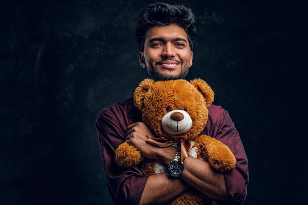 Usmívající se mladý Indián ve stylové tričko objímá medvídka a pohled z boku. - Fotografie, Obrázek
