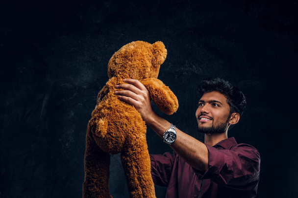 Indian guy in stylish shirt looks at his lovely teddy bear while holding it in hands. Studio photo against a dark textured wall - Photo, Image
