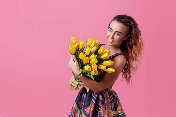 Cheerful young lady in stylish fashionable dress being excited to get bouquet of spring yellow flowers tulips on women's day isolated over pink background.  - Photo, image