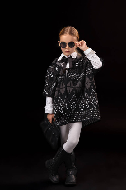 Bambina con occhiali neri, poncho scuro, stivali neri e una pochette in mano
 - Foto, immagini