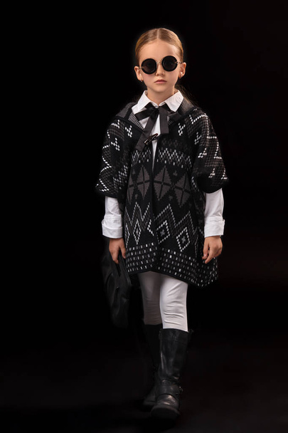 Pieni tyttö mustissa laseissa, tumma poncho, mustat saappaat ja kytkin kädessään
 - Valokuva, kuva