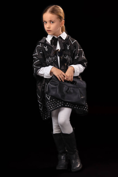 Little girl in a dark poncho, black boots and a clutch in her hand - Фото, зображення
