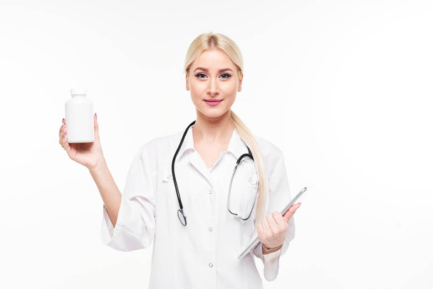 Attractive young female doctor with stethoscope pointing at white pill bottle isolated on white background - Foto, Imagen
