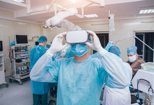 Doctor in a surgical room with virtual reality glasses on the background of the real operation. Modern technologies - Фото, изображение