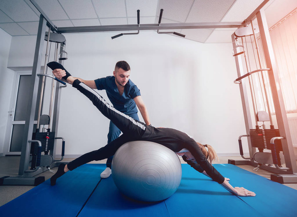 Concept of rehabilitation . Beautiful young woman doing exercises with ball under supervision of physiotherapist. Exercise therapy - Zdjęcie, obraz