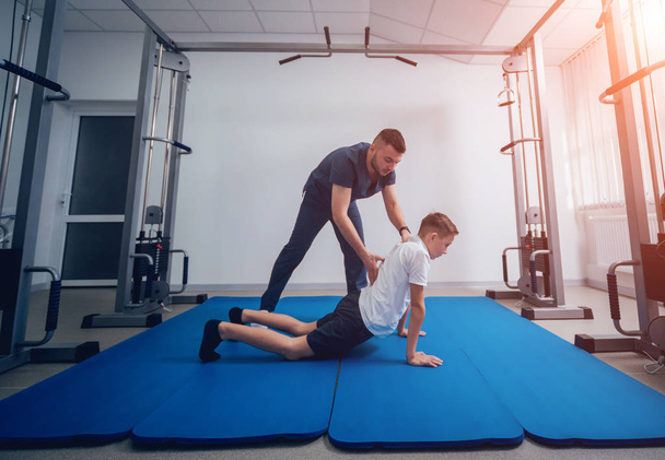 Concept of rehabilitation. Young boy doing exercises on mat under supervision of physiotherapist. Exercise therapy - Foto, Imagen