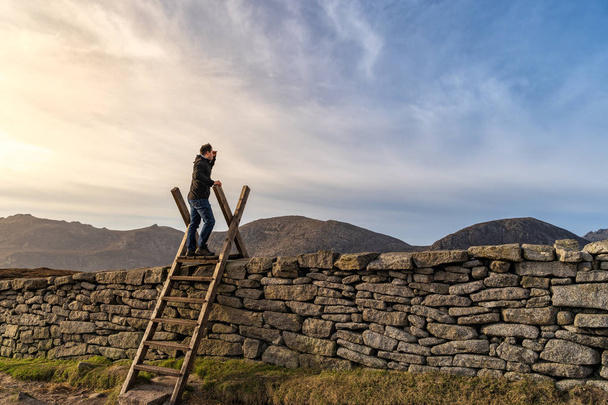 Чоловік стоїть на драбині на кам'яній стіні в горах з заходом сонця на задньому плані
 - Фото, зображення