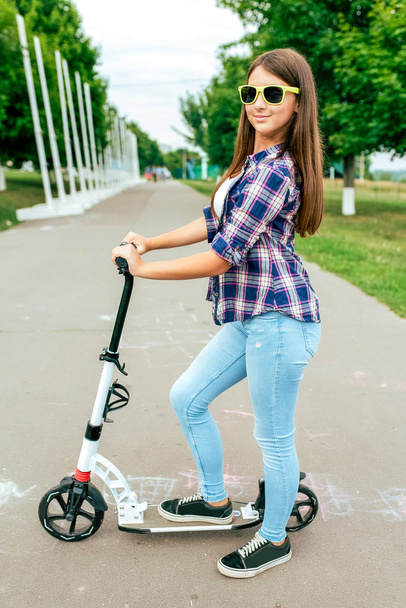 Menina adolescente estudante 10-15 anos. Em uma scooter no parque da cidade de verão. Feliz sorrindo em óculos de sol. Descansando depois da escola montando uma scooter no fim de semana. Em roupas casuais, camisa e jeans em tênis
. - Foto, Imagem