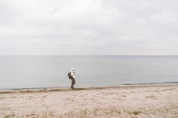 Un hombre caucásico joven turista caucásico con una mochila negra en una playa de arena cerca del mar Báltico en invierno. Viaje temático solo. Pensamientos y sueños con vistas al horizonte
 - Foto, Imagen