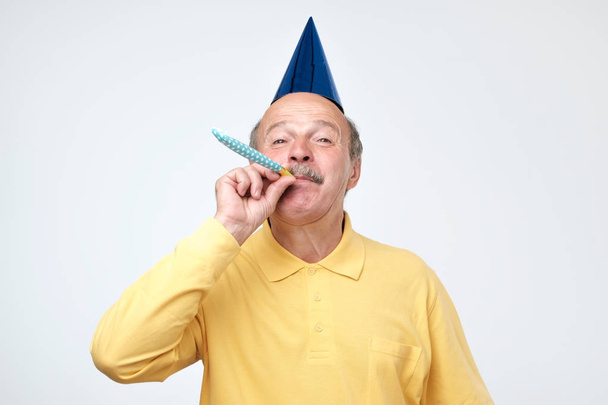 Starší muž foukání strany roh a zírala na kamery s vzrušený pohled zároveň slaví narozeniny - Fotografie, Obrázek