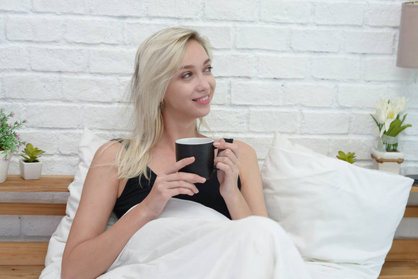 Bonita jovem sorrindo mulher sentada na cama com uma xícara de café
 - Foto, Imagem